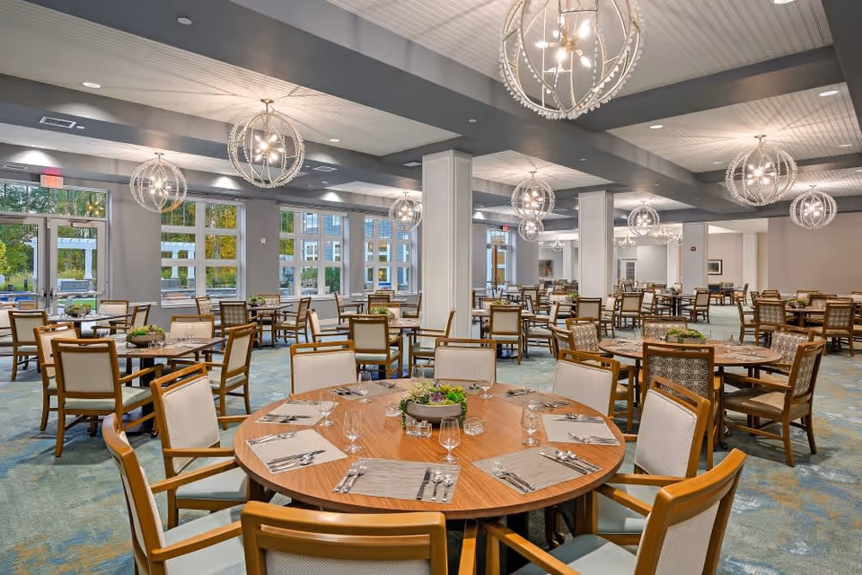 A spacious dining room with multiple round and square wooden tables surrounded by cushioned chairs. Each table is set with placemats, silverware, glasses, and a centerpiece of greenery. Large windows line the walls, allowing natural light to fill the room. Modern spherical chandeliers hang from the ceiling, and the carpet has a subtle blue and green pattern.