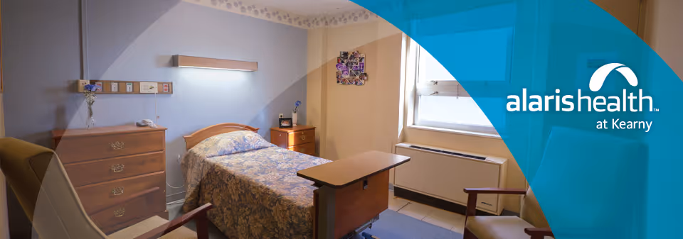 A small, neatly arranged bedroom in a senior living facility with a single bed covered in a floral bedspread, a wooden dresser, a bedside table with a lamp, a chair, and a window letting in natural light. The room has light-colored walls and a simple, cozy atmosphere.