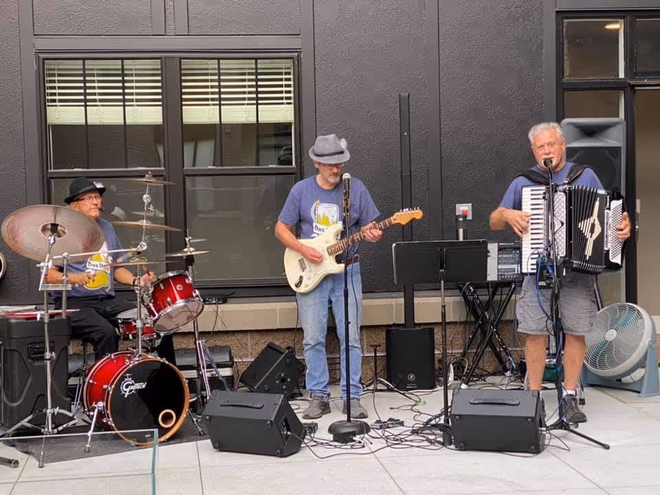 Three elderly men performing music outdoors in front of a building. One man is playing a drum set, another is playing an electric guitar, and the third is playing an accordion and singing into a microphone. They are standing on a paved area with speakers and musical equipment around them.