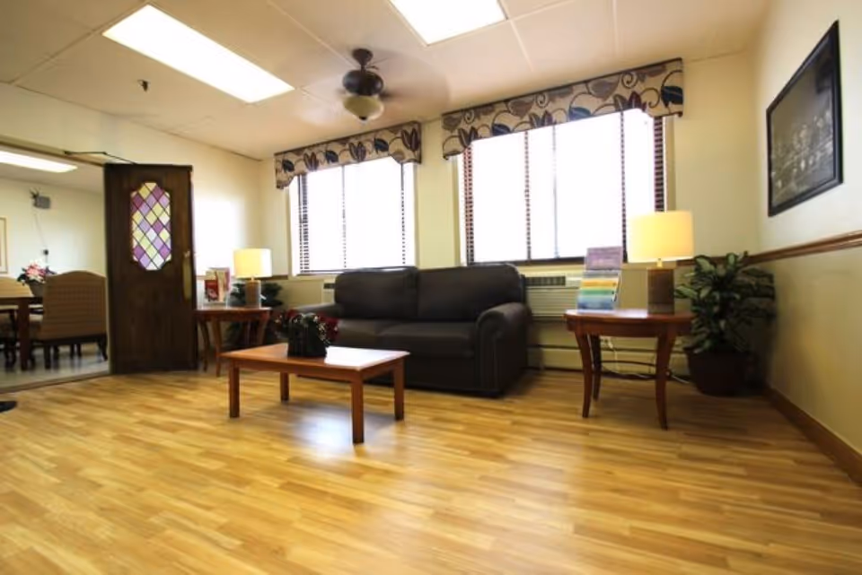 A reception-style lounge with a dark sofa, coffee table, side tables with lamps, and large windows over wood flooring.