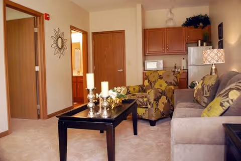 Cozy living room with a sofa, patterned armchair, coffee table with candles, and a small kitchenette in the background.