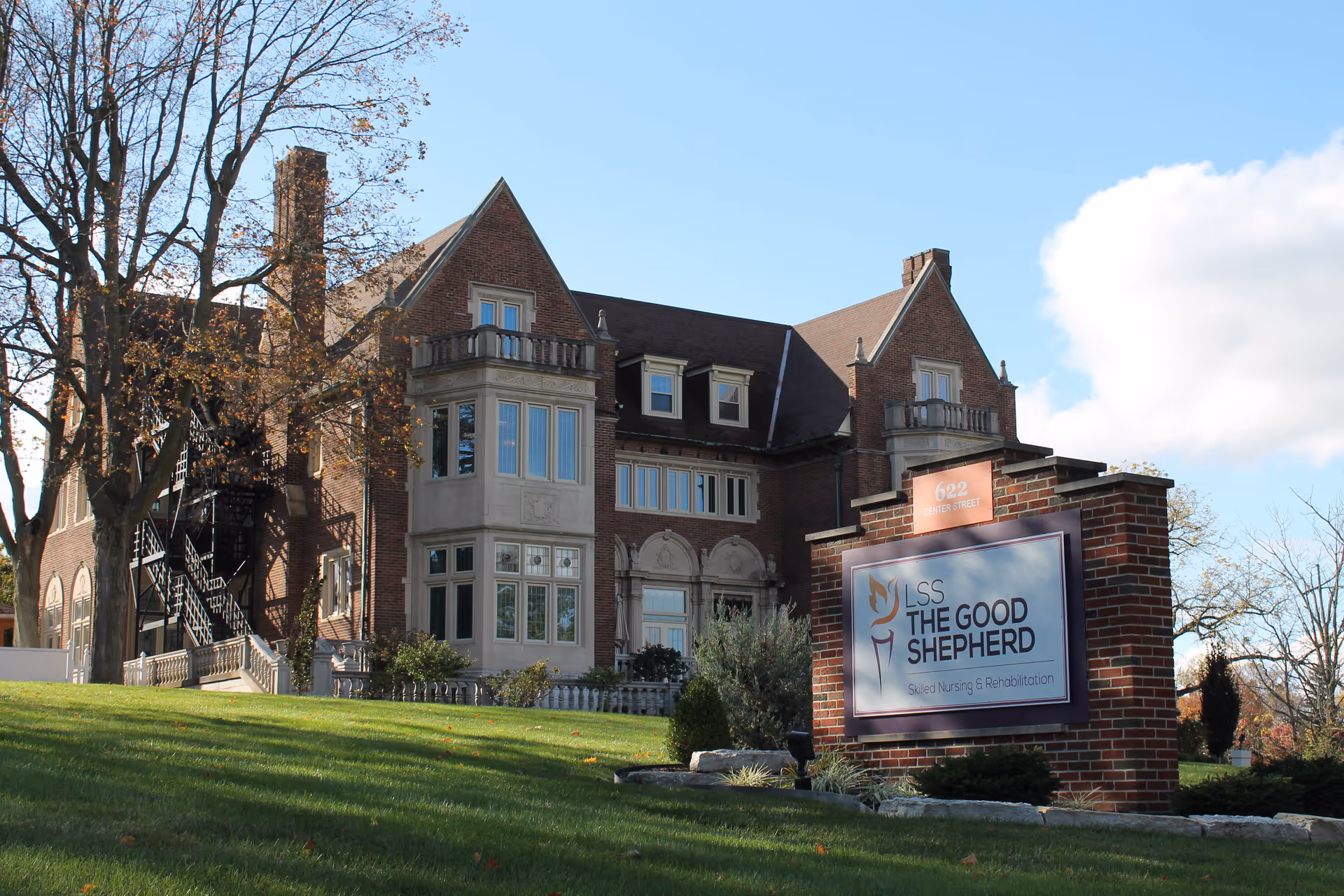 Exterior view of a large brick building with multiple windows and a steep roof, surrounded by trees and a well-maintained lawn. In front of the building is a brick sign that reads 'LSS THE GOOD SHEPHERD Skilled Nursing & Rehabilitation' with the address '622 Scherers Street' displayed above it. The sky is clear with some clouds.