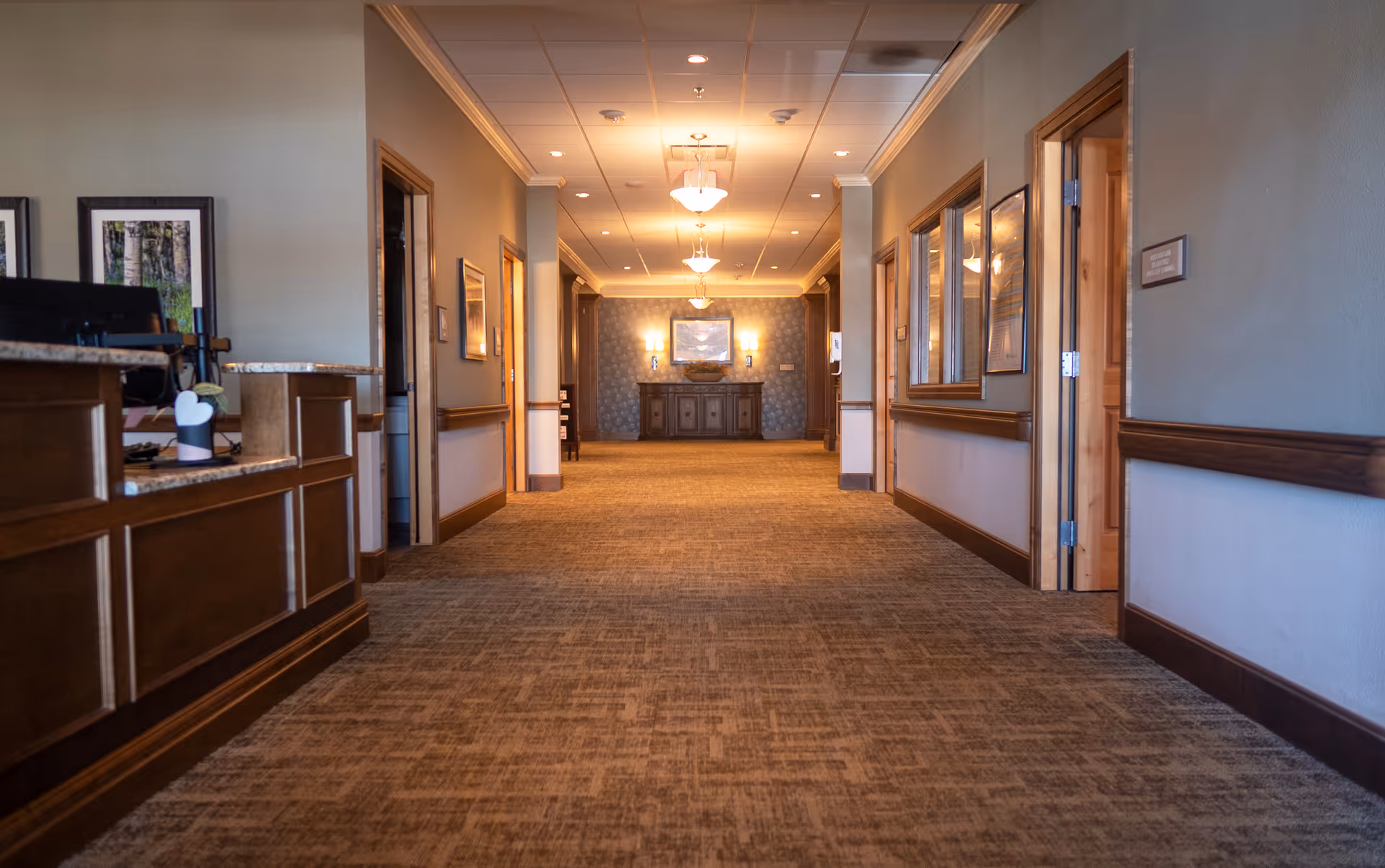 A wide hallway in a senior living facility with carpeted floors, wooden trim, and soft overhead lighting. On the left side, there is a reception desk with a computer and decorative items. The hallway has several doorways and framed pictures on the walls, leading to other rooms. At the far end, there is a wooden cabinet with a framed picture above it and wall sconces on either side.