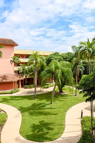 Outdoor courtyard area at Elison Independent Living of Lake Worth featuring a green lawn with several palm trees, curved concrete walkways, and a multi-story building with a red-tiled roof and peach-colored walls under a partly cloudy blue sky.