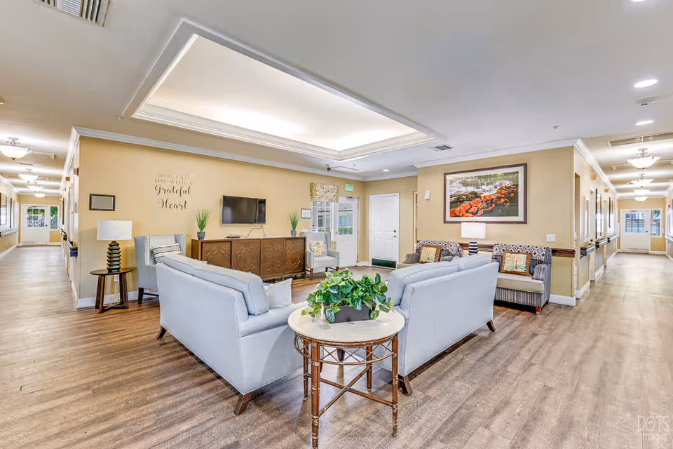 A bright and spacious senior living facility common area with light blue sofas arranged around a small round table with a green plant. The room has wooden flooring, beige walls, and a ceiling light fixture. There is a TV mounted on the wall above a wooden cabinet, two armchairs, and a framed picture of flowers on the wall. Hallways extend on both sides of the room with doors and windows visible.