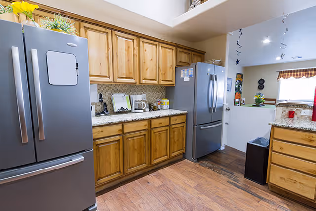 A kitchen area with wooden cabinets, granite countertops, two stainless steel refrigerators, a coffee maker, and various kitchen items on the counter. The floor is wooden, and there is a small trash bin near the counter. The kitchen opens into a dining area with a window and some decorations hanging from the ceiling.