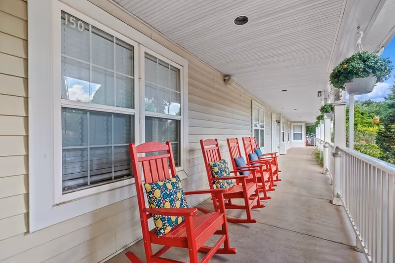 A long covered porch with a row of red rocking chairs, each with a colorful cushion. The porch has white railings and hanging green plants, with beige siding on the building and windows along the wall.