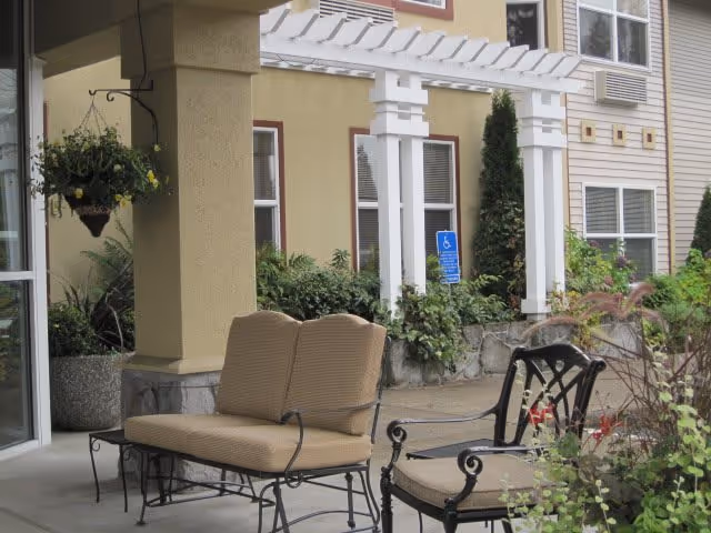 Outdoor seating area at a senior living facility with cushioned chairs and a small table under a covered patio. There is a hanging planter with flowers, greenery along the building wall, and a white pergola structure near the windows. A blue handicap parking sign is visible in the background.