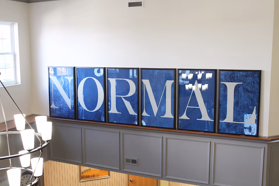 Interior common area showing six framed blue panels spelling 'NORMAL' mounted above a gray-paneled railing with a chandelier and a nearby window.