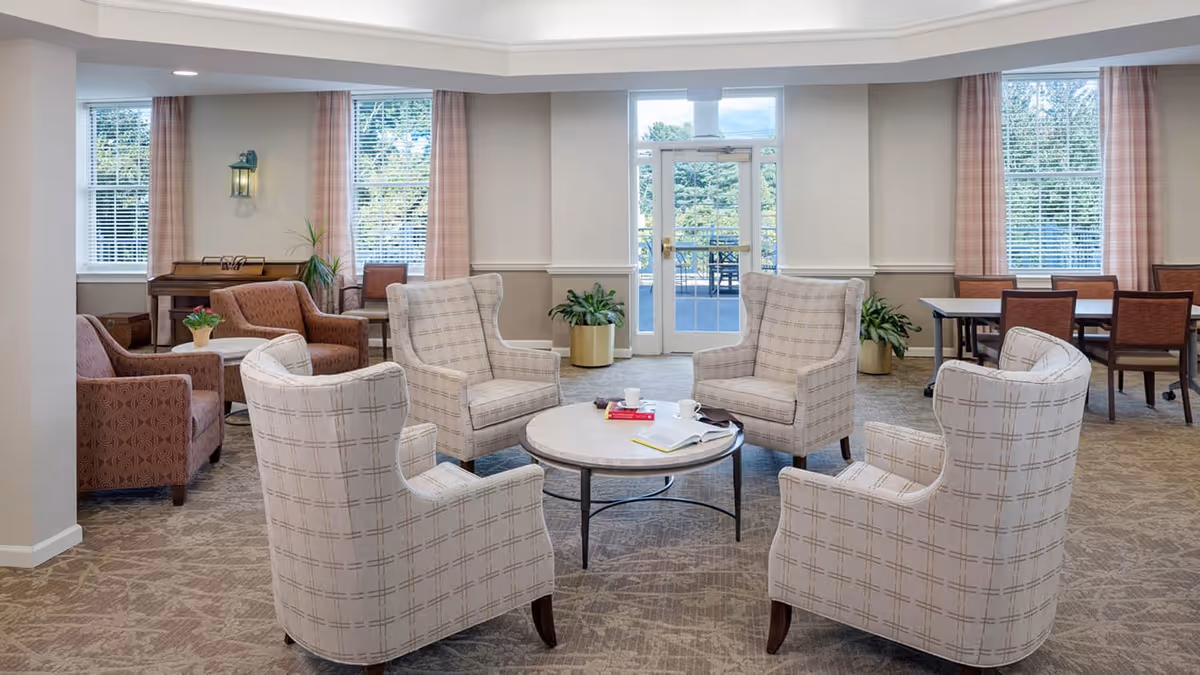 A bright and spacious living room area with four beige patterned armchairs arranged around a round coffee table holding books and cups. In the background, there are additional seating areas with brown armchairs, a piano, and a dining table with chairs near large windows with pink curtains. A glass door leads to an outdoor patio with greenery visible outside.
