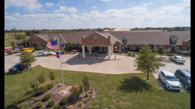 Front exterior view of Legend Oaks Healthcare and Rehabilitation - Ennis, showing a single-story brick building with a covered entrance, a circular driveway, several parked cars, an American flag on a flagpole, and landscaped greenery under a partly cloudy sky.