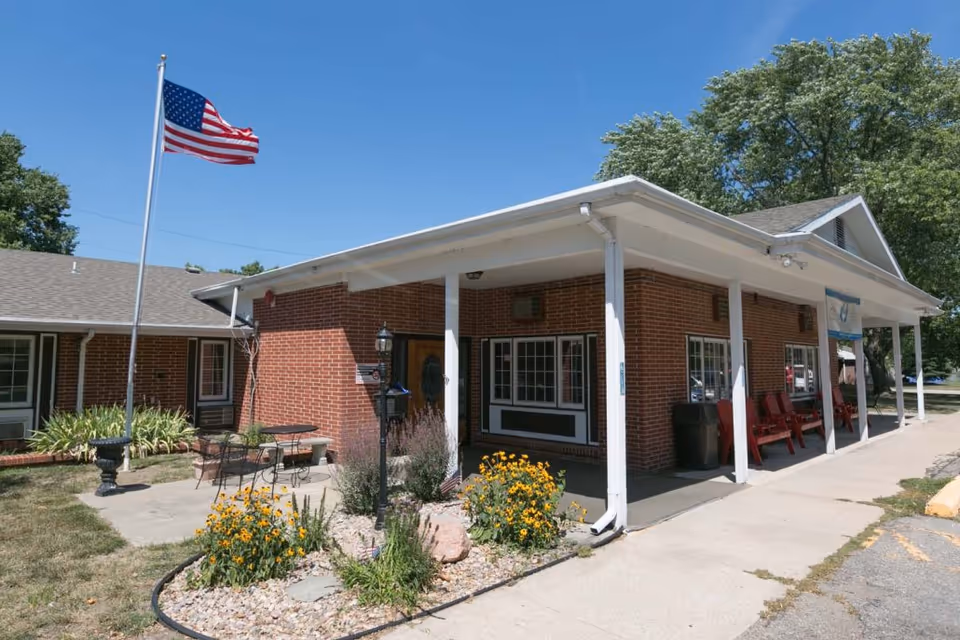 Exterior view of a single-story brick building with a covered porch supported by white columns. There are red chairs on the porch, a small garden with yellow flowers in front, and an American flag on a flagpole to the left. The sky is clear and blue.