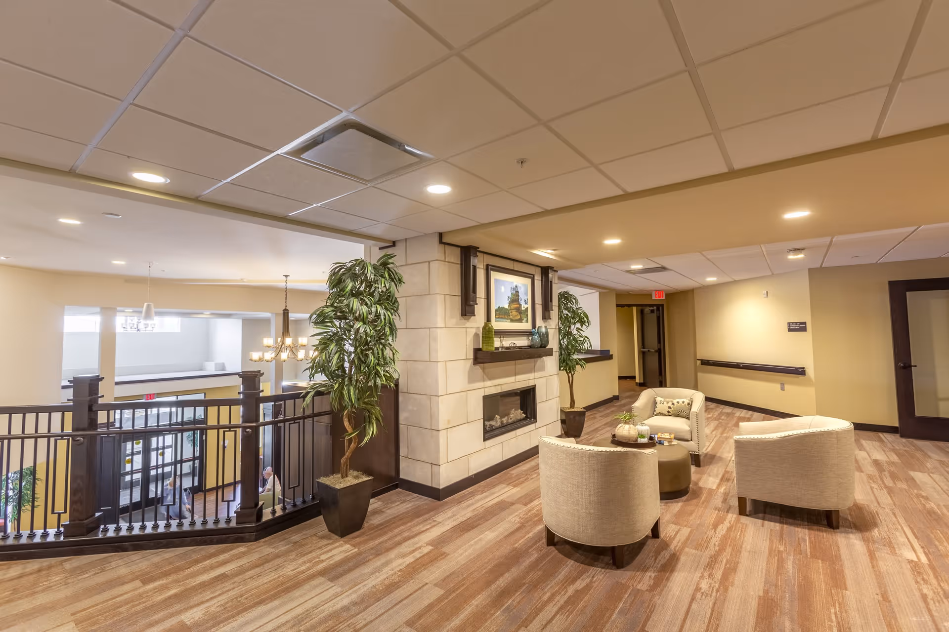 A cozy seating area in a senior living facility with three beige armchairs arranged around a small round table. The space features a modern fireplace with decorative vases and a framed picture above it. There are two large potted plants on either side of the fireplace. The area overlooks a lower level with large windows and a chandelier, and the flooring is wood-patterned. The walls are painted in warm neutral tones with recessed lighting in the ceiling.