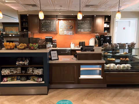 Interior view of a cafe counter at Phoebe Berks with baskets of fruit and snacks on the left, a sign that says 'Order Here', a glass display case with baked goods on the right, and three hanging pendant lights above. Behind the counter are wooden cabinets and chalkboard menus.
