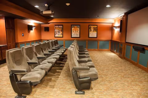 A small theater room with two rows of cushioned reclining seats facing a large projection screen. The walls are painted orange and teal with wooden trim, and three framed movie posters are displayed on the back wall. The room has dim lighting with wall sconces and a projector mounted on the ceiling.