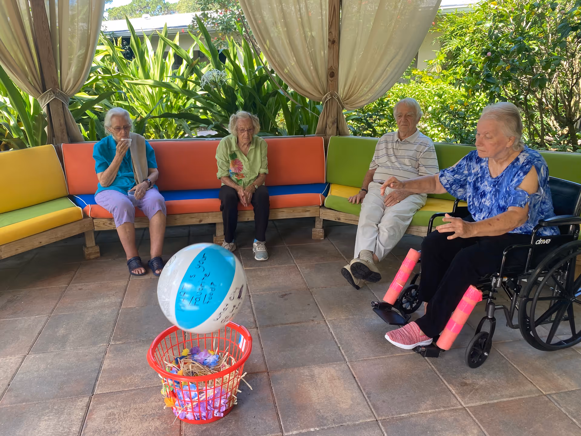 Four elderly individuals sitting on colorful cushioned benches and a wheelchair under a covered outdoor patio area, playing a game where a beach ball is tossed towards a red basket filled with decorative items.