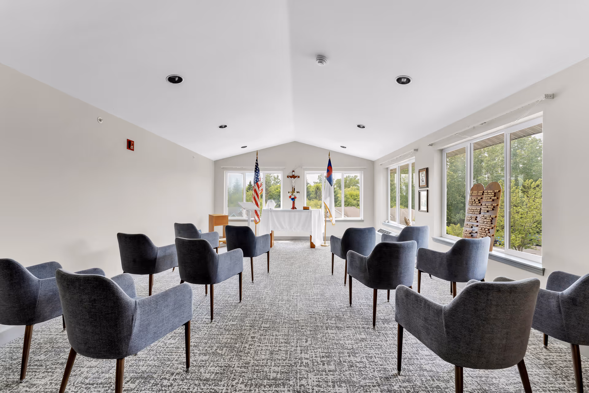 A small chapel or meditation room with rows of gray upholstered chairs facing a white altar table at the front. The altar has two flags behind it, one American and one Christian, with a cross and a flower arrangement on top. Large windows on the right side let in natural light and show green trees outside. The room has a light gray carpet and white walls with recessed ceiling lights.
