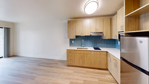A modern kitchen area with light wood cabinets, a white countertop, a stainless steel refrigerator, and a blue tiled backsplash. The kitchen is adjacent to an empty room with light wood flooring and a sliding glass door leading outside.