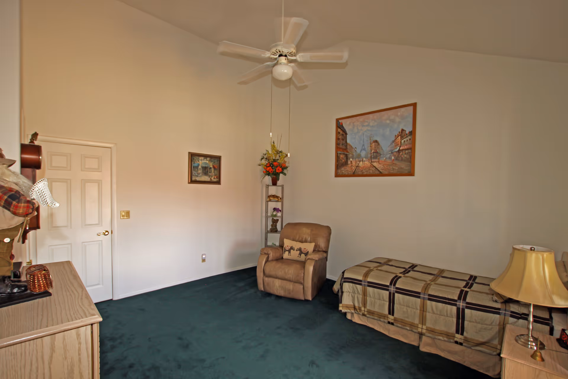 A bedroom with a single bed covered in a plaid bedspread, a beige recliner chair with a decorative pillow, a wooden dresser, a small side table with a lamp, two framed paintings on the walls, a ceiling fan with a light, and a green carpeted floor.