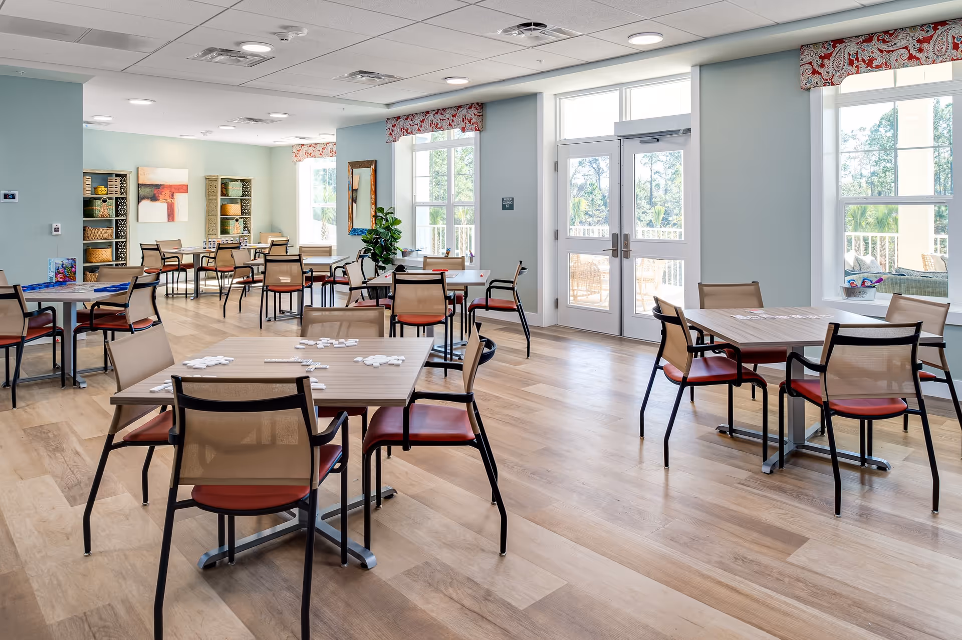 Bright communal activity and dining room with multiple tables and chairs, large windows, and a door leading outside.