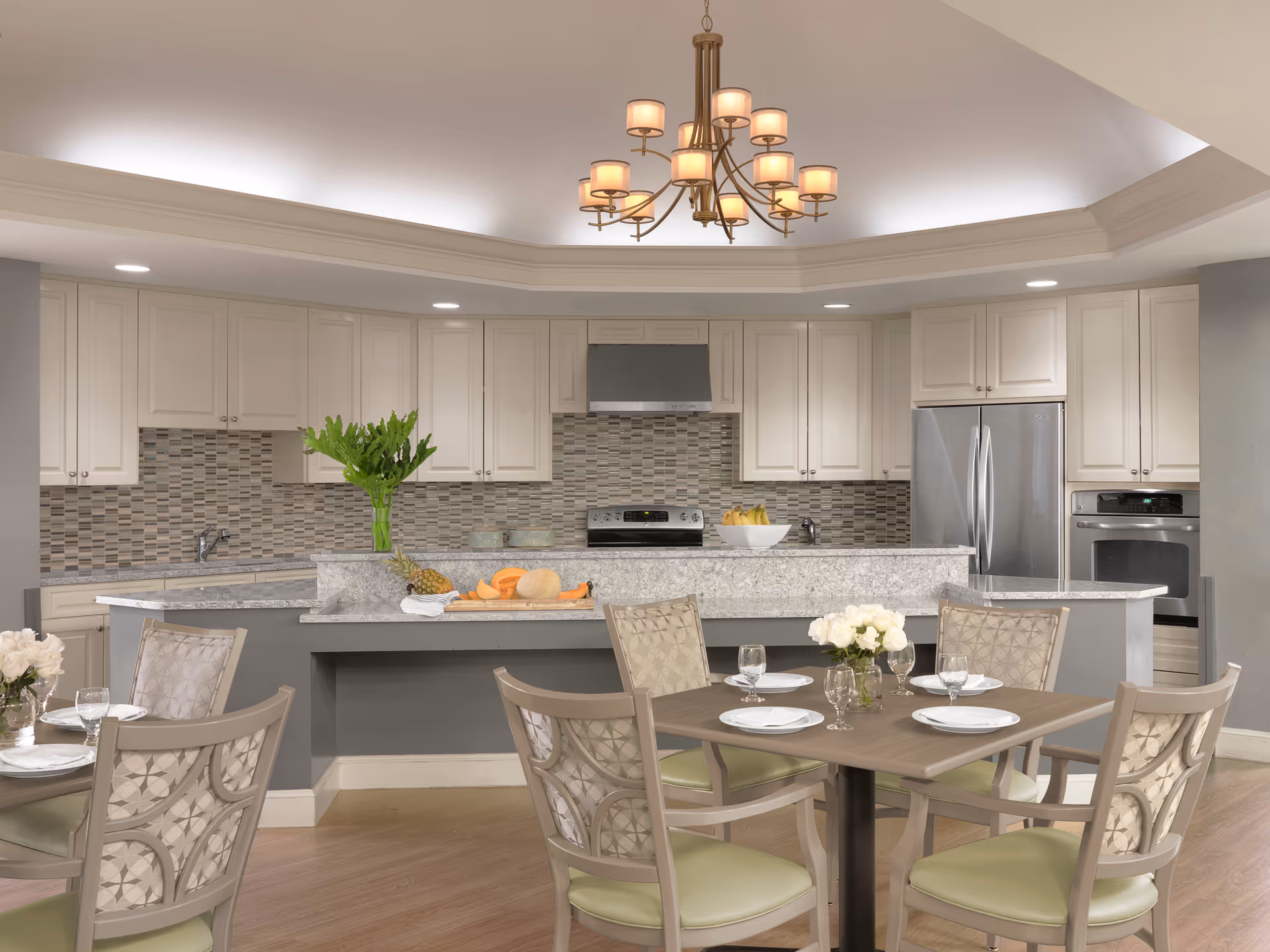 A modern kitchen and dining area with a large island countertop in the center, white cabinets, stainless steel appliances including a refrigerator, oven, and stove with a hood. The backsplash features a mosaic tile design. In front of the kitchen island, there is a dining table set with four chairs, each with light green cushions and patterned backs. The table is set with white plates, glasses, and a small vase of white flowers. A chandelier with multiple lights hangs from the ceiling above the kitchen area.