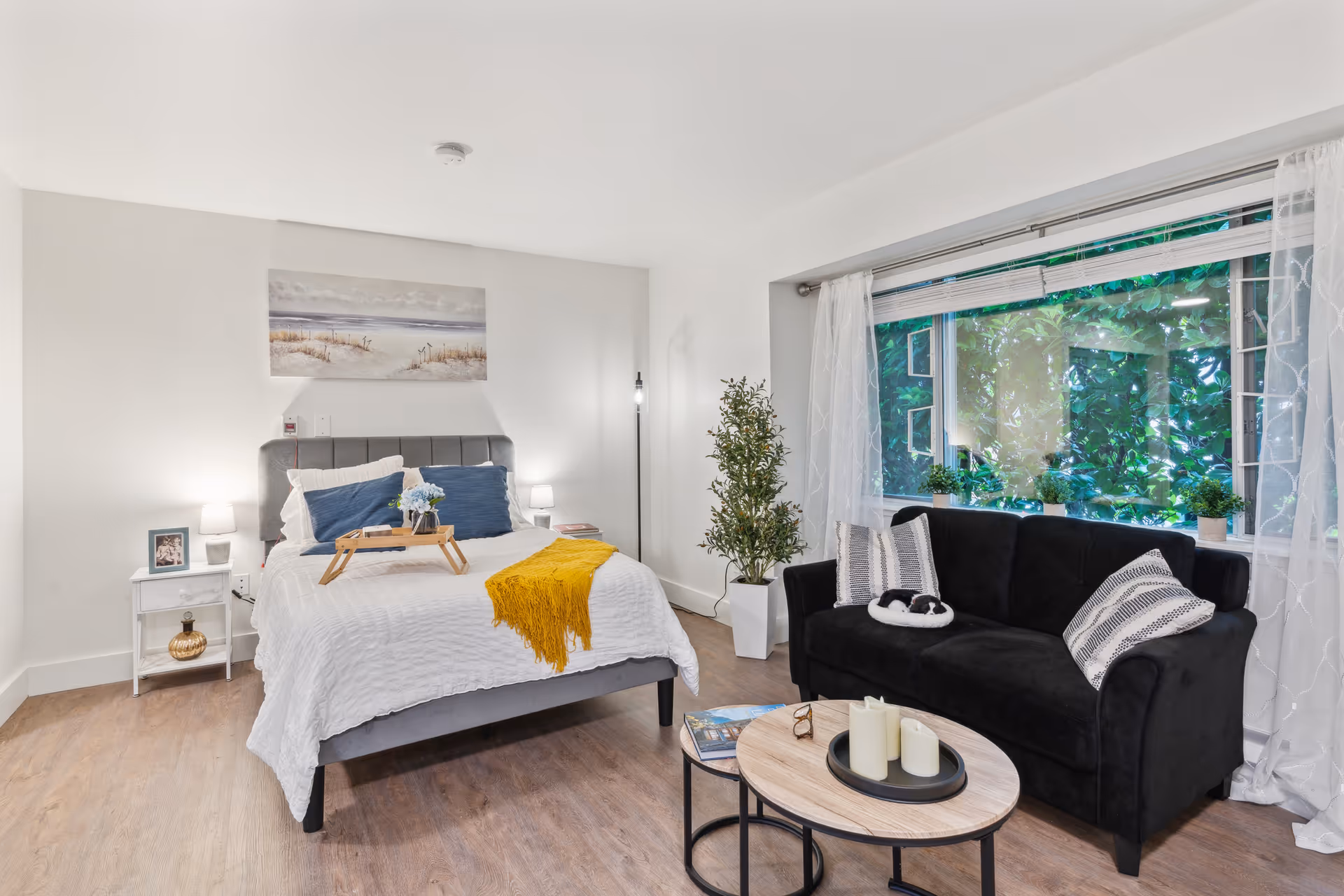 A bright and cozy bedroom featuring a neatly made bed with white bedding, blue pillows, and a yellow throw blanket. A wooden tray with flowers is placed on the bed. To the right, there is a black sofa with patterned pillows and a small black and white cat curled up on it. In front of the sofa, there are two round wooden coffee tables with candles and a book. A large window with sheer white curtains lets in natural light and shows greenery outside. The room has wooden flooring, a white nightstand with a framed photo and lamp, a tall potted plant, and a floor lamp in the corner.