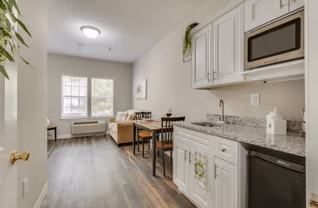 Interior view of a small apartment or studio with a kitchenette on the right featuring white cabinets, a granite countertop, a sink, a microwave, and a mini refrigerator. In the background, there is a dining table with four chairs and a beige sofa near two large windows with blinds. The floor is wood-style laminate and the walls are painted light beige.