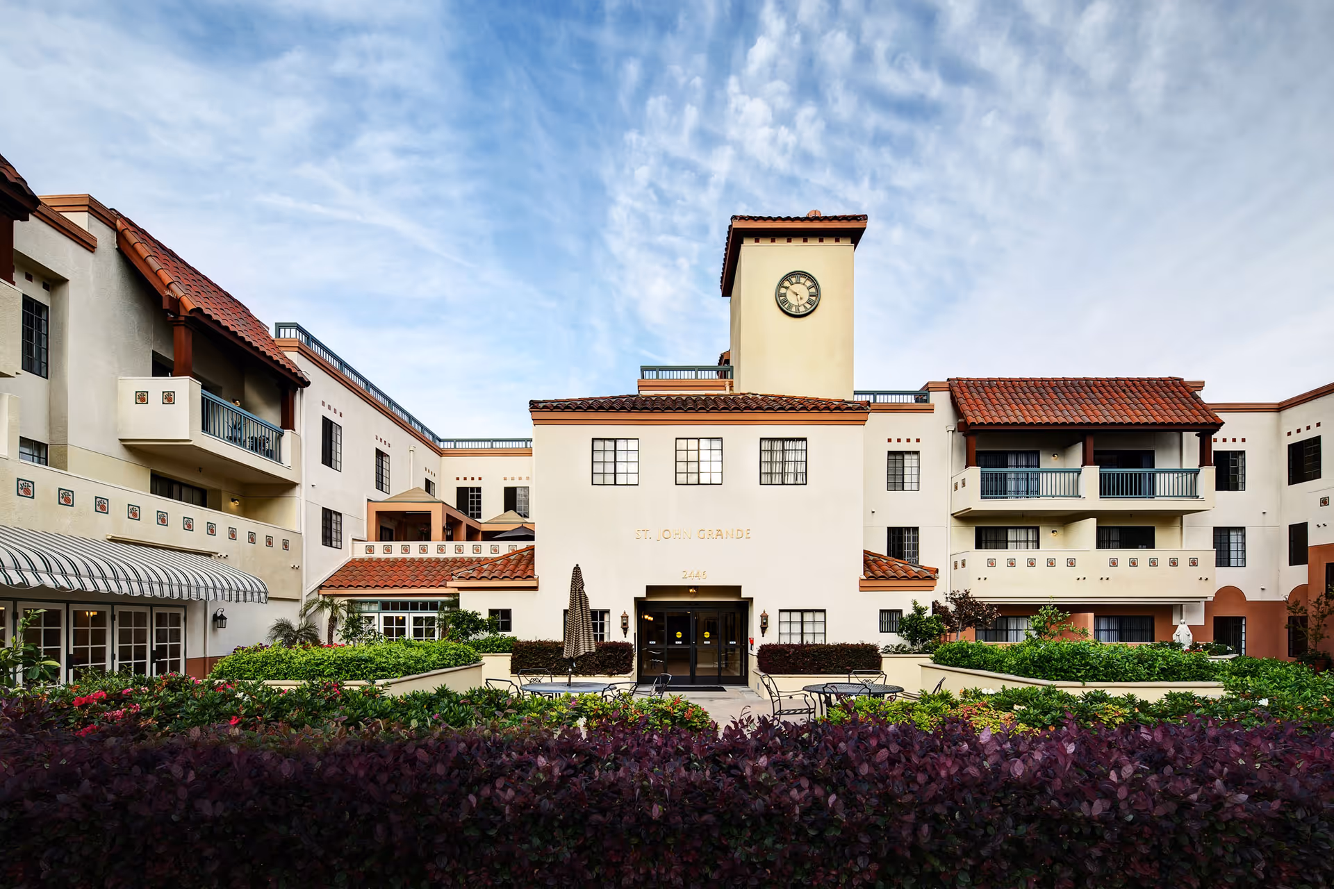 Exterior front view of St. John Grande retirement and care center building with a clock tower, balconies, and a landscaped garden area with tables and chairs in front.