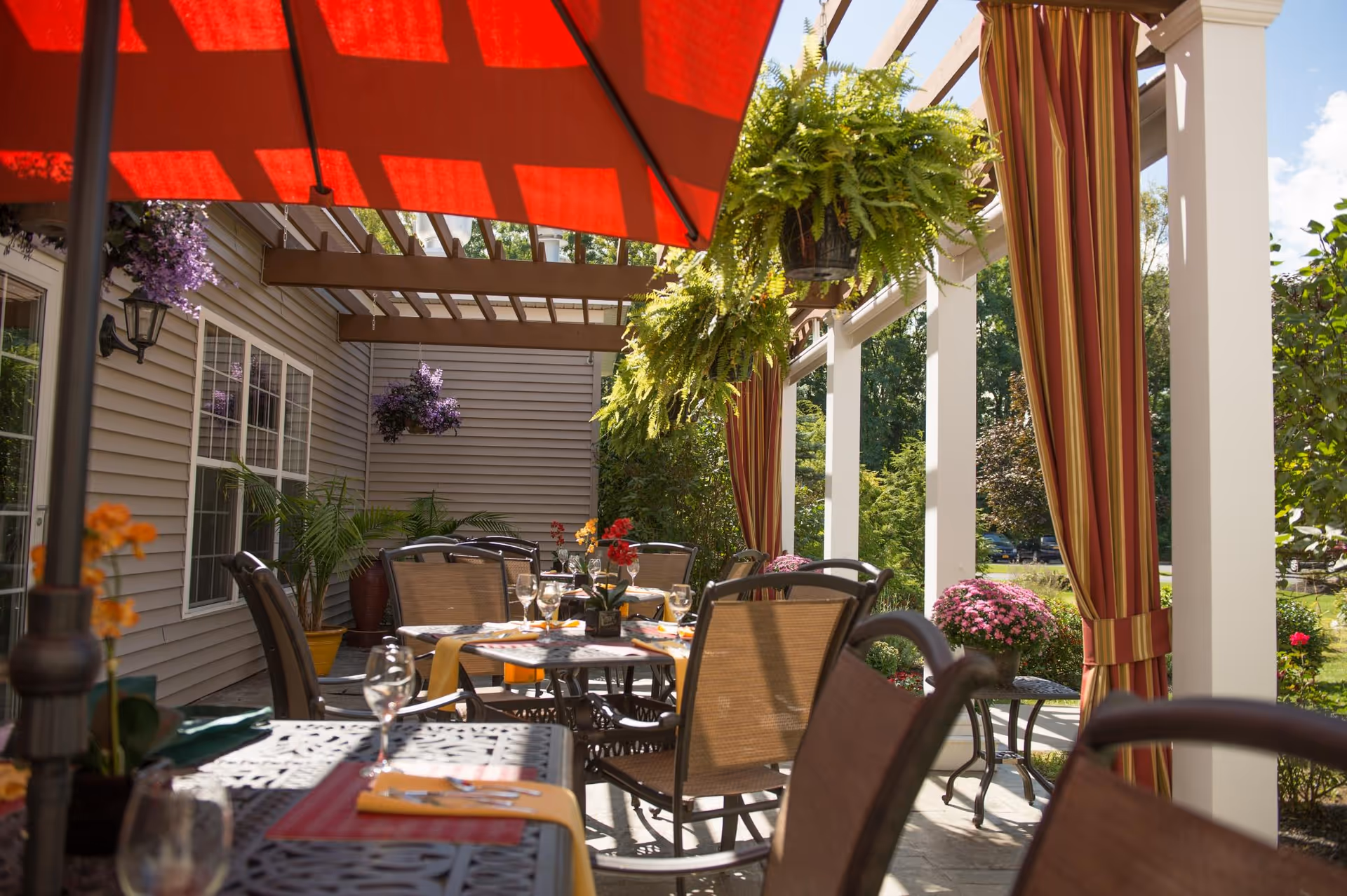 Outdoor patio dining area with metal tables and chairs under a pergola with hanging plants and red-striped curtains. A large red umbrella provides shade, and the area is decorated with flowers and greenery.