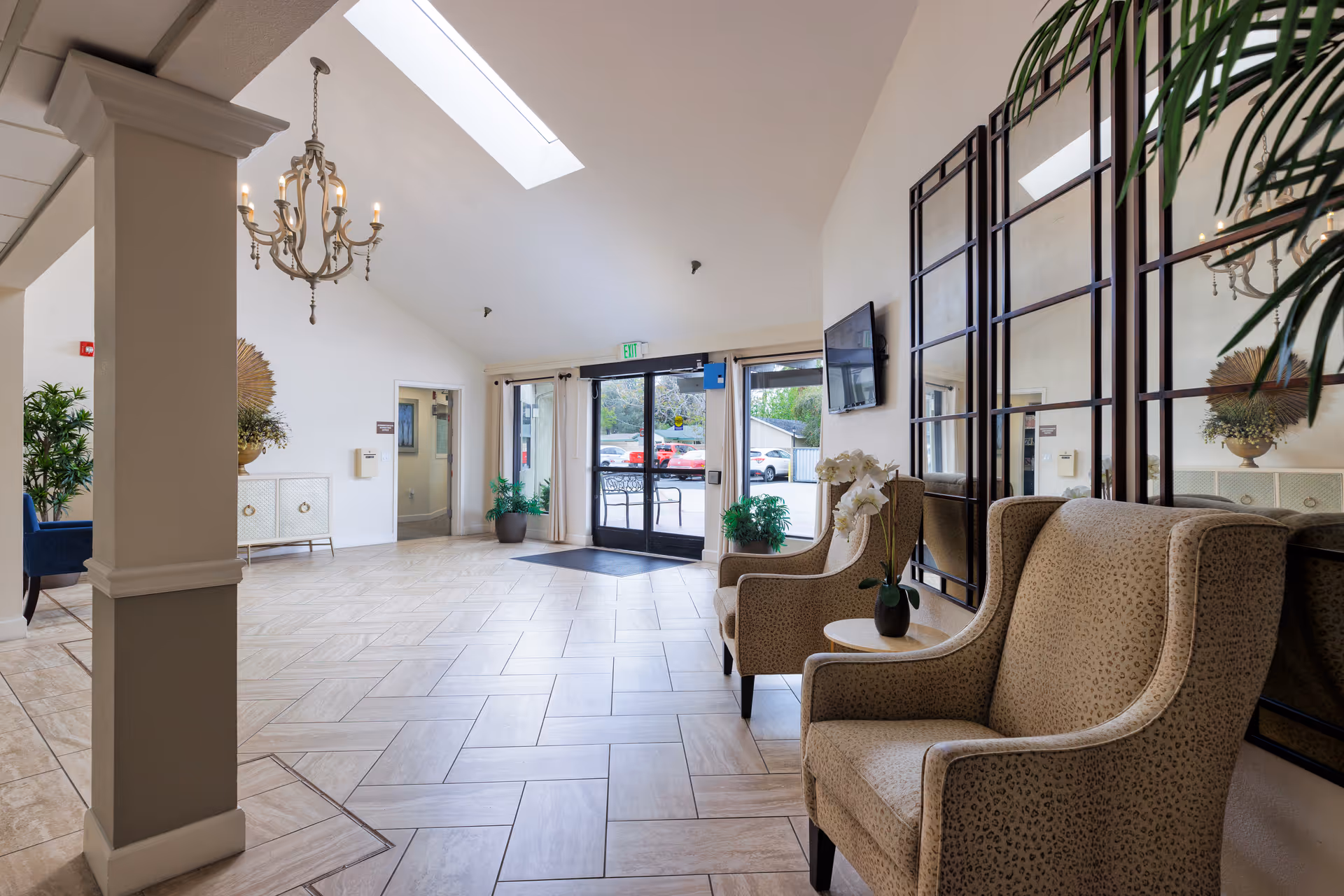 Bright lobby with tiled floor, upholstered armchairs and decorative mirrors facing glass entrance doors under a skylight.