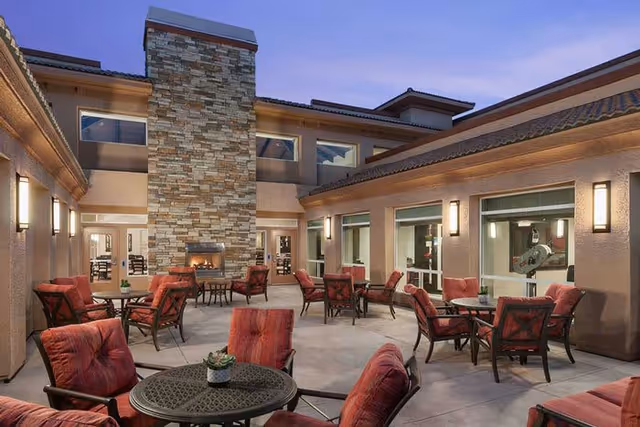 Outdoor patio area at a senior living facility with multiple round tables and cushioned chairs arranged around a central stone fireplace. The area is surrounded by the building with windows and wall-mounted lights, under a twilight sky.