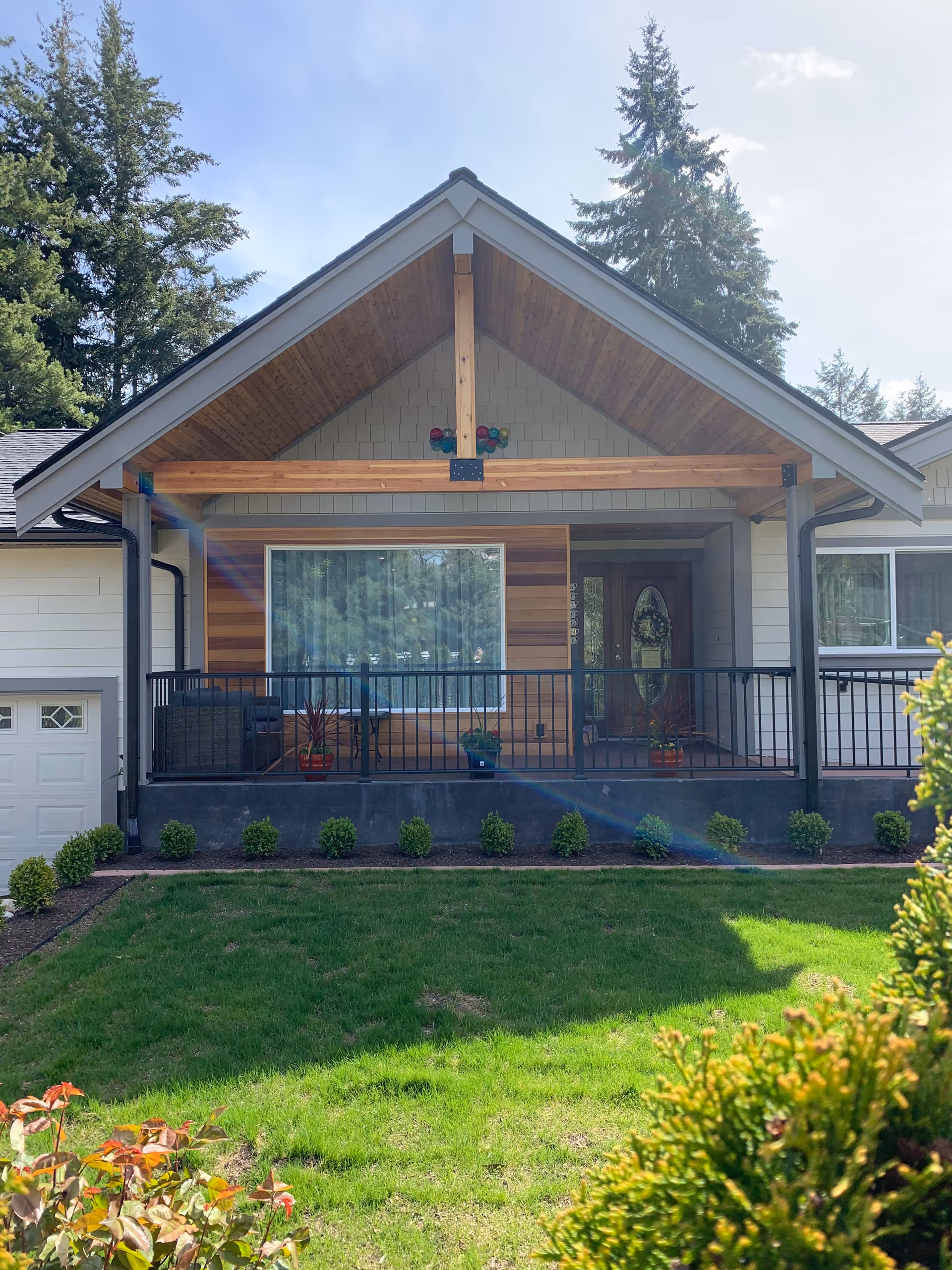Front exterior view of a single-story house with a covered porch featuring wooden beams, a large window with curtains, a glass front door, and a black metal railing. The yard in front has green grass, small bushes, and some plants. Tall trees are visible in the background under a clear sky.