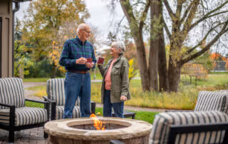 An elderly man and woman standing outdoors near a fire pit, holding cups and smiling at each other. They are surrounded by cushioned patio chairs and trees with autumn foliage in the background.