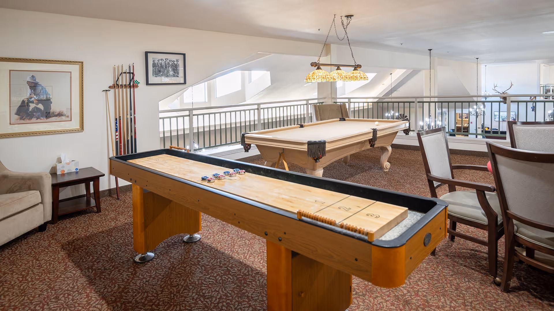 A recreational room with a shuffleboard table in the foreground and a billiards table in the background. The room has carpeted flooring, several chairs, a small side table with tissues, and framed artwork on the walls. The space is well-lit with natural light coming through large windows and hanging light fixtures above the billiards table.