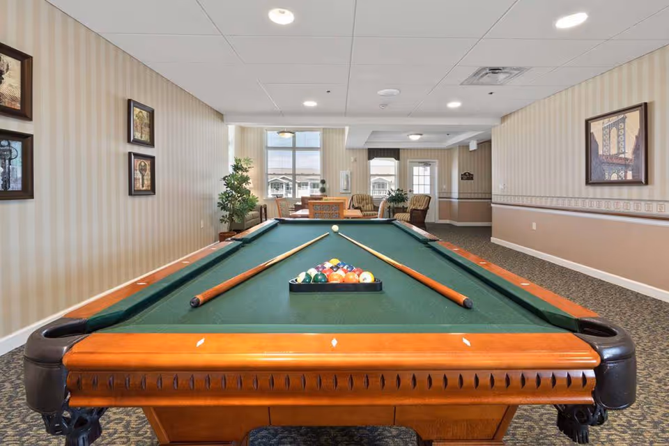 Interior view of a recreational room featuring a pool table with two cues and a rack of billiard balls set up for a game. The room has beige striped wallpaper, framed artwork on the walls, carpeted floor, and a seating area with chairs and a table near large windows letting in natural light.