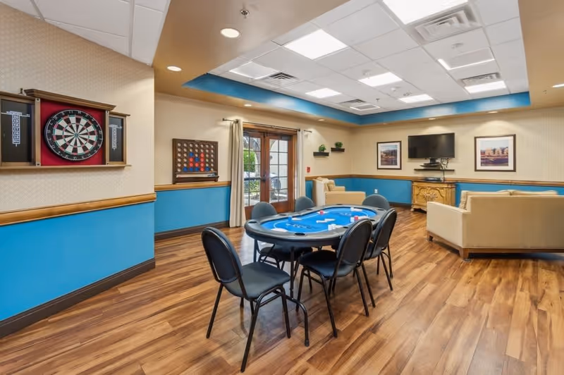 A recreational room with a poker table surrounded by six chairs in the center. The room has wood flooring, blue and beige walls, and a ceiling with recessed lighting. On one wall, there is a dartboard and a large Connect Four game. The opposite wall features a flat-screen TV mounted above a wooden cabinet, flanked by two framed pictures. Two beige sofas are arranged near the TV, and a glass door with curtains leads outside.