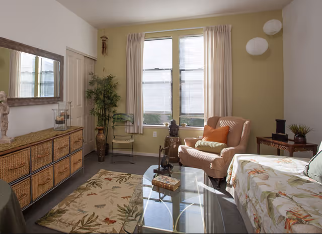 Sunlit living room with an armchair, glass coffee table, wicker storage cabinet, and a daybed by a window.