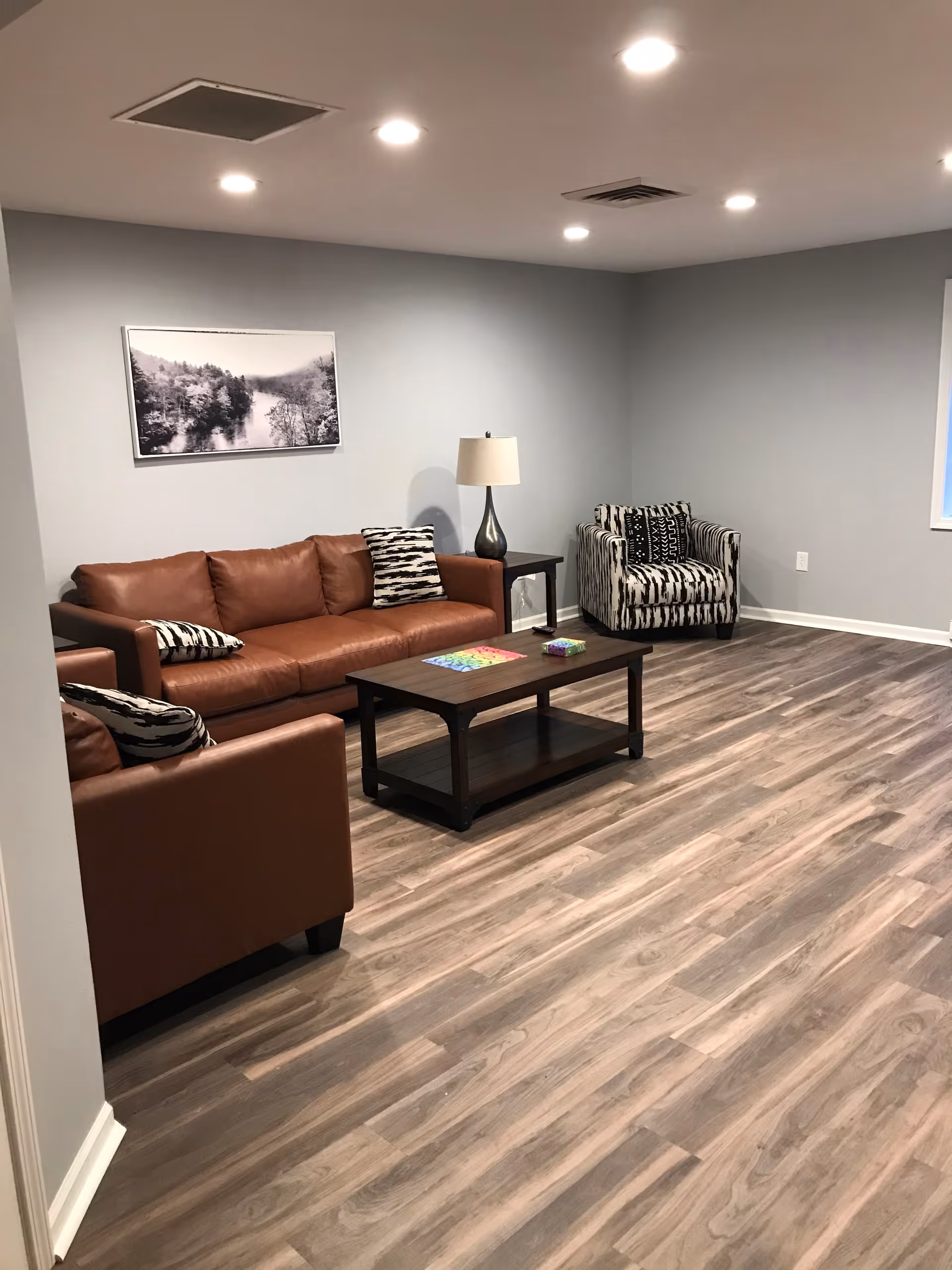 A modern living room with a brown leather sofa, a matching armchair, and a patterned armchair. There is a dark wooden coffee table with colorful items on it, a side table with a lamp, and a black and white framed photograph of a river and trees on the light gray wall. The floor is wood laminate and the ceiling has recessed lighting.