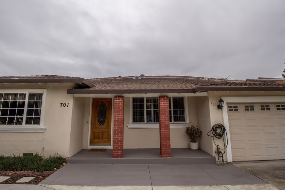 Front exterior view of a single-story house with a wooden front door, two red brick columns supporting a small porch roof, a window with white framing, and an attached garage with a closed door. The house number 701 is visible on both sides of the entrance. The sky is overcast.