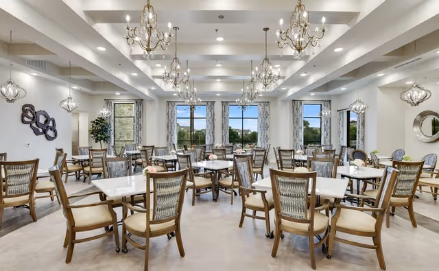 A spacious and elegant dining room with multiple square tables and wooden chairs with cushioned seats. The room features large windows with patterned curtains allowing natural light to enter, several ornate chandeliers hanging from the ceiling, and decorative wall mirrors and art pieces. The floor is tiled, and the overall ambiance is bright and inviting.