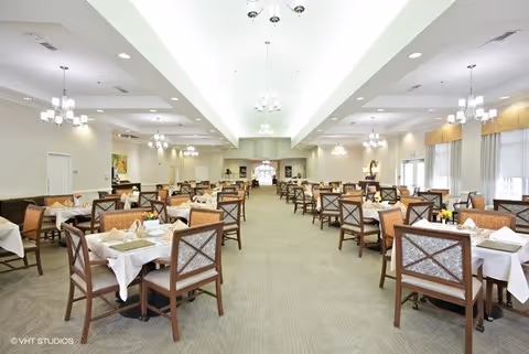 A spacious dining room with multiple tables covered with white tablecloths, each set with placemats, napkins, and small flower arrangements. The room has beige carpeting, soft lighting from chandeliers, and large windows with beige curtains allowing natural light to enter.