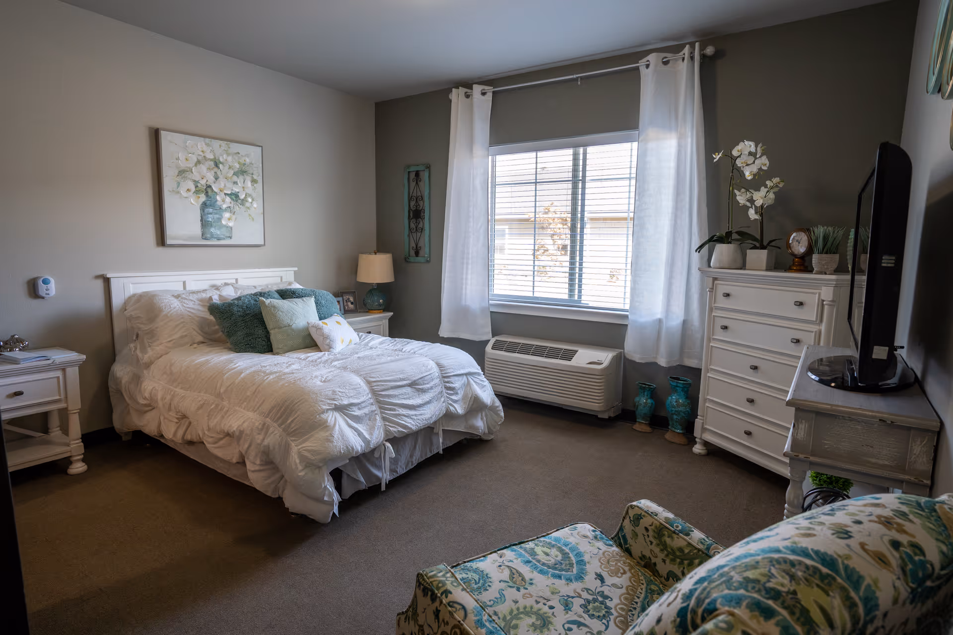 A cozy bedroom with a white bed adorned with multiple pillows, a nightstand with a lamp, a window with white curtains, a white dresser with decorative items including a clock and plants, a patterned armchair, and a wall-mounted TV.
