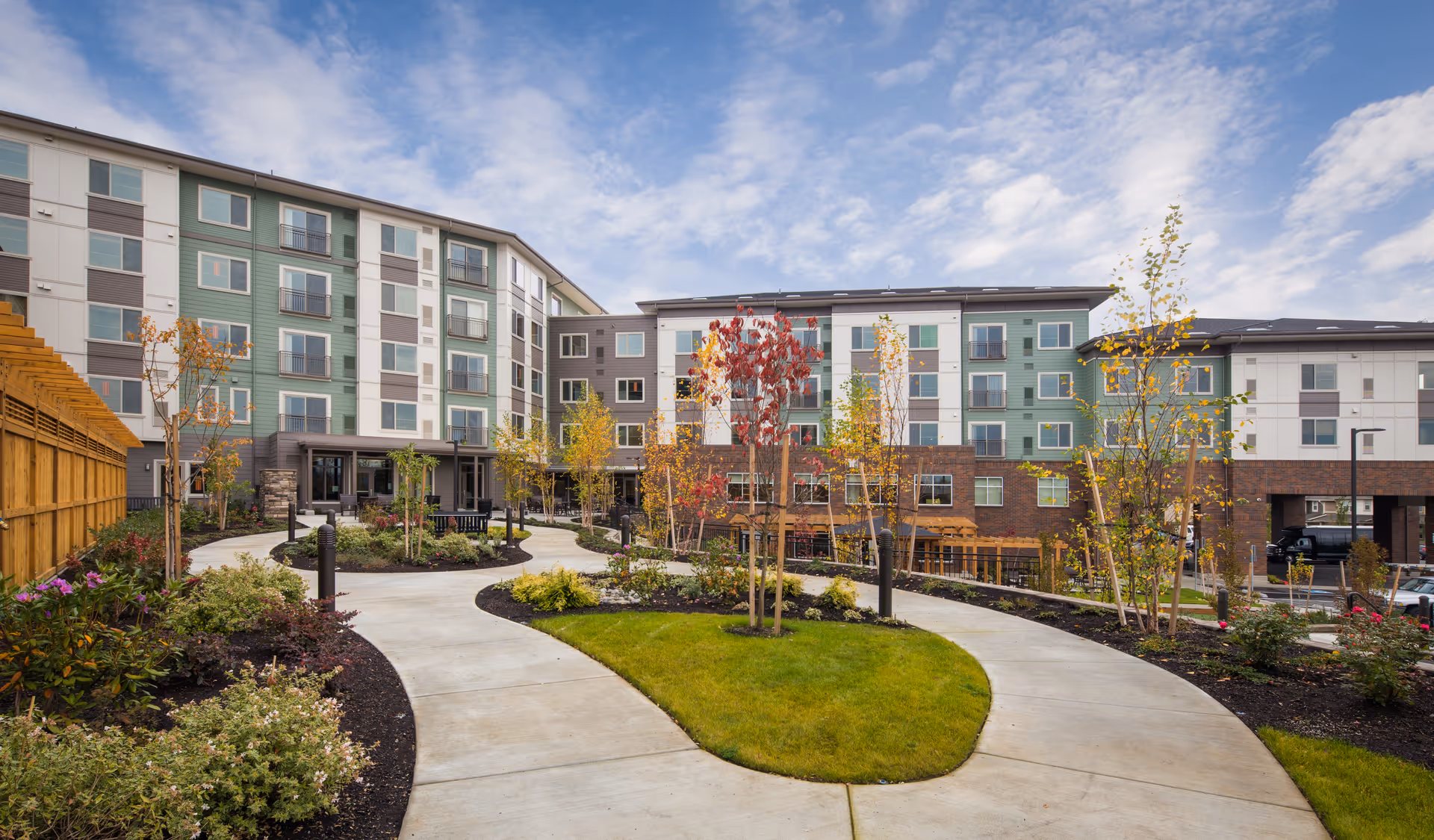 Outdoor view of a multi-story senior living facility building with a landscaped garden area featuring a curved concrete pathway, young trees, shrubs, and green grass under a partly cloudy sky.