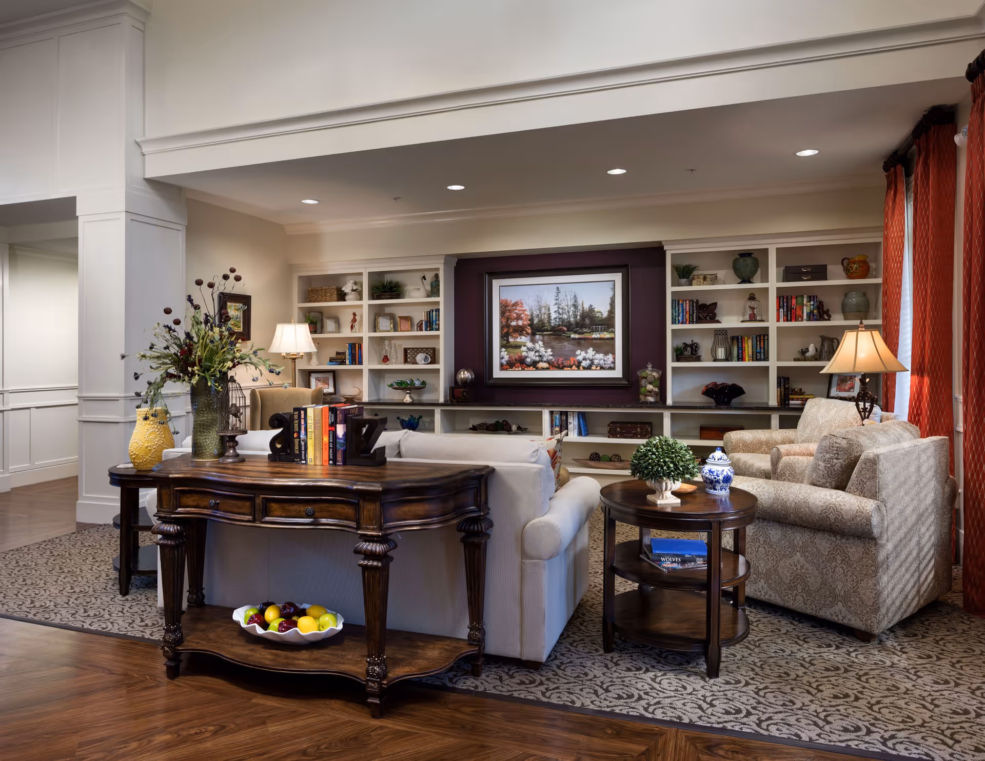 A cozy living room area with a white sofa and patterned armchair arranged around a wooden coffee table and side table. Behind the sofa is a wooden console table with decorative items and books. The back wall features built-in white shelves filled with books, plants, and decorative objects, with a framed painting of a scenic landscape in the center. The room has warm lighting from table lamps and natural light coming through red curtains on the right.