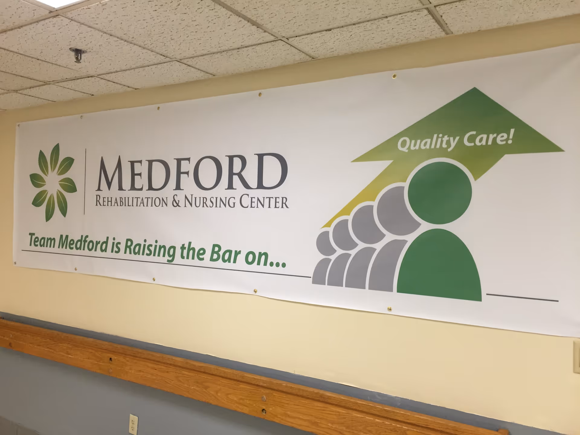 A banner hanging on a beige wall inside a building. The banner reads 'MEDFORD Rehabilitation & Nursing Center' with a green flower-like logo on the left. Below it says 'Team Medford is Raising the Bar on...' and to the right is a graphic of five stylized human figures ascending with a green arrow above them that says 'Quality Care!'