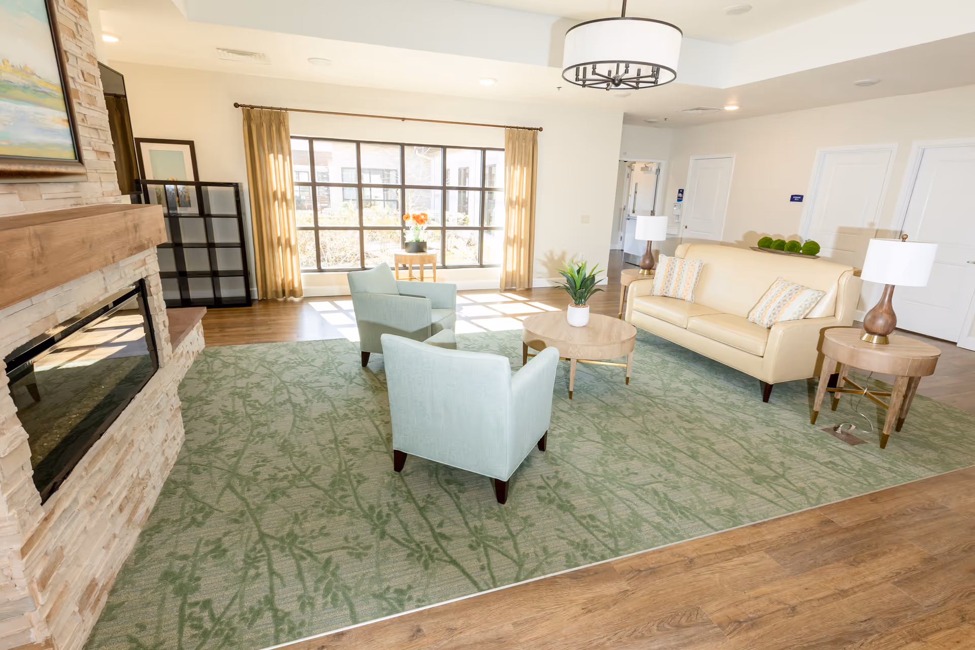 A bright and spacious living room with a large window letting in natural light. The room features a beige leather sofa with patterned pillows, two light blue armchairs, a round wooden coffee table with a small plant, and two matching side tables with lamps. There is a stone fireplace on the left side and a green patterned rug covering part of the wooden floor.