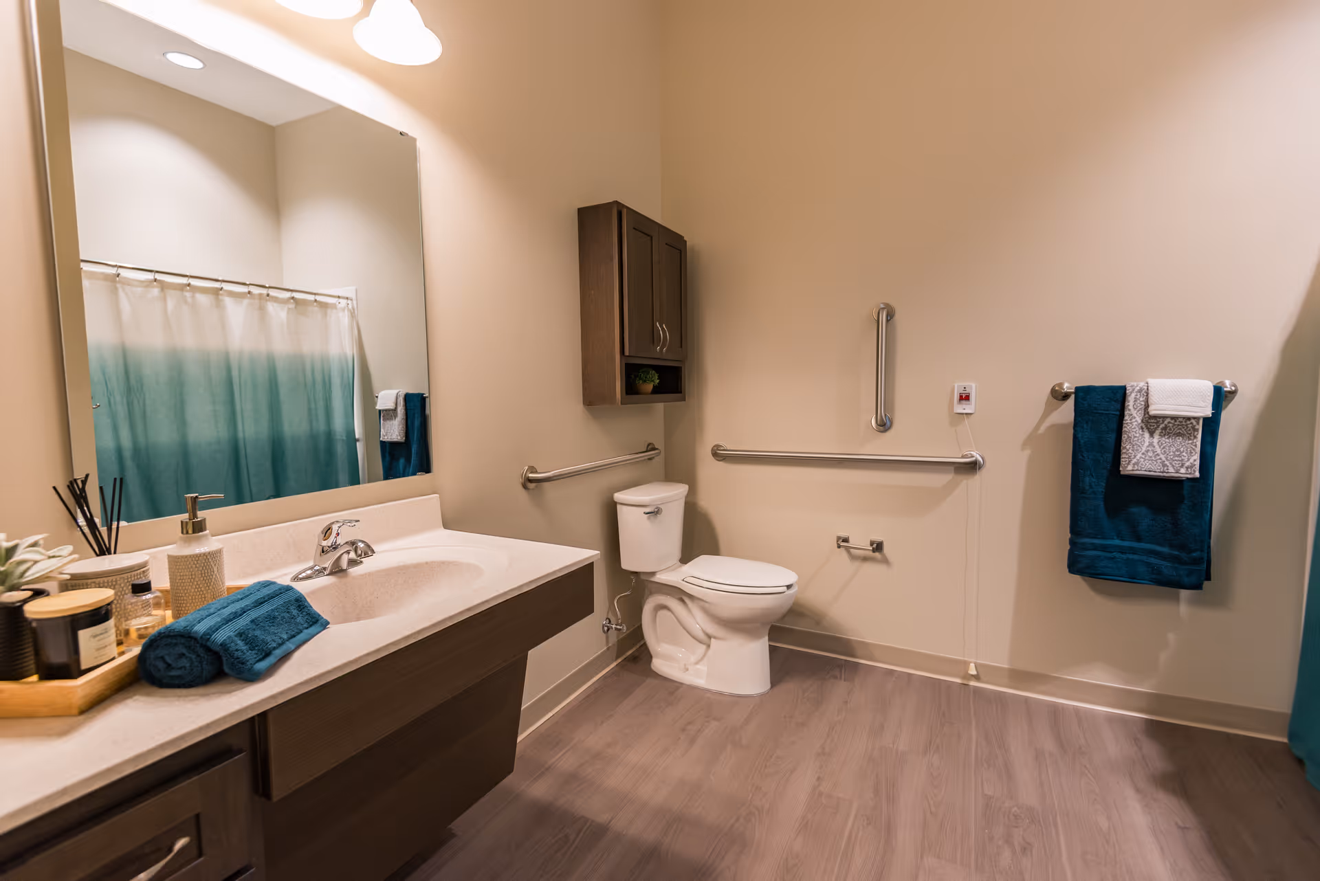 A spacious senior living bathroom with a large mirror above a sink countertop. The bathroom features a toilet with grab bars on the walls for accessibility, a towel rack with folded towels, a shower curtain with a gradient blue design, and wooden cabinetry. The floor is covered with wood-like flooring and the walls are painted a light beige color.