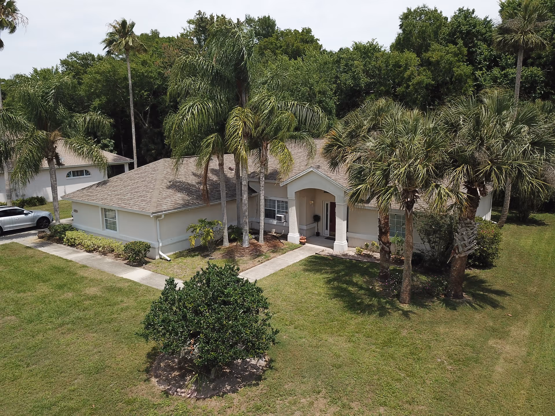 A single-story residential building with a light-colored exterior and a shingled roof, surrounded by palm trees and green grass. A concrete pathway leads to the front entrance, which has a small covered porch. There is a car parked on the left side near the building, and dense trees are visible in the background.