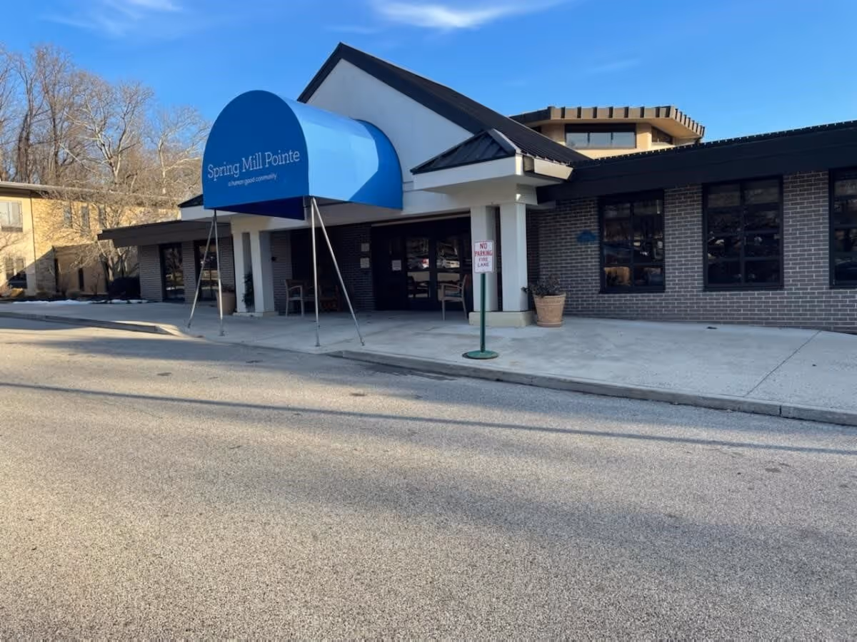 Exterior front entrance of Spring Mill Pointe facility with a blue awning displaying the facility name. The building has brick walls and large windows, with a sidewalk and a 'No Parking Fire Lane' sign in front.