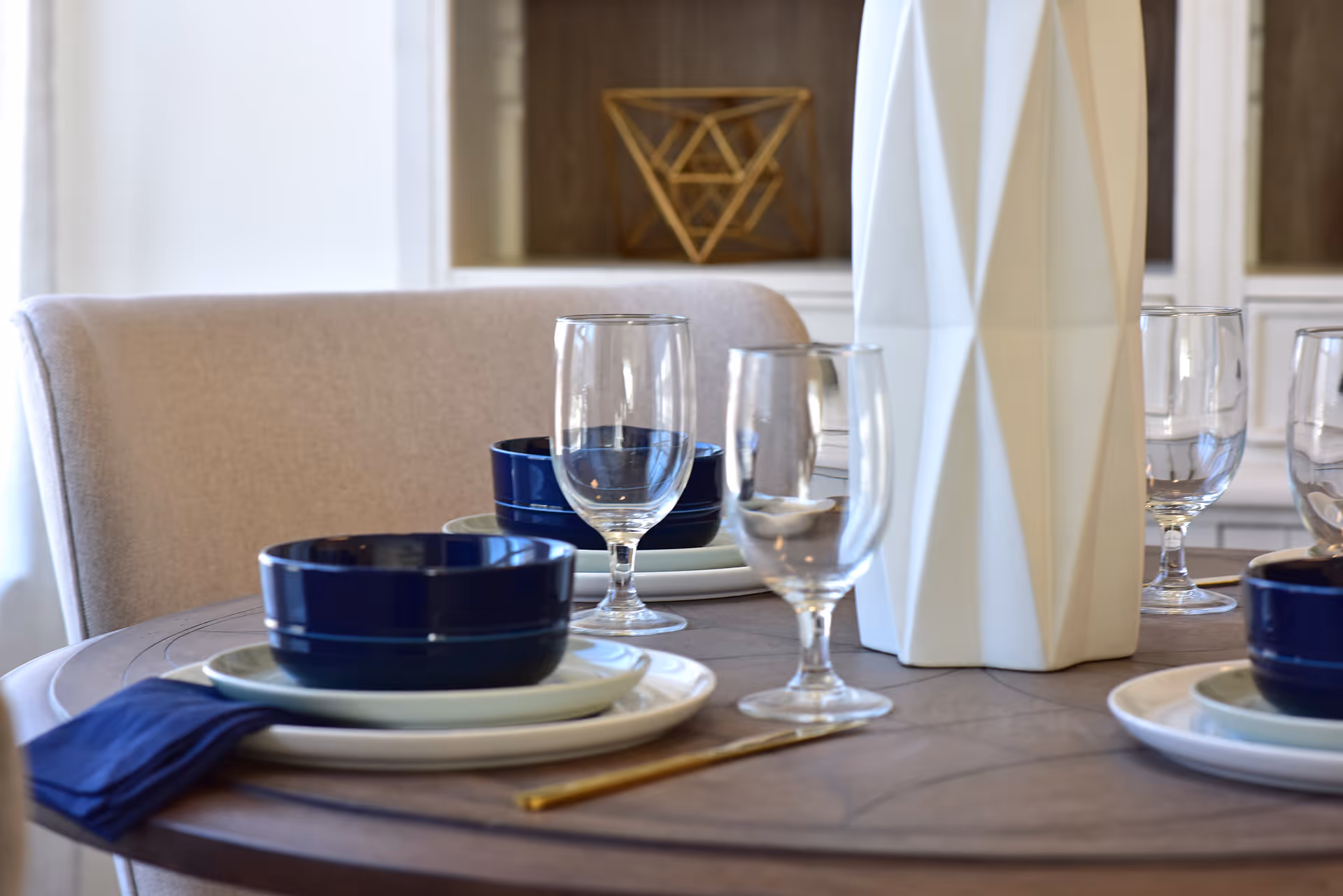 Close-up of a dining table set with plates, blue bowls, wine glasses, and a tall white geometric vase.